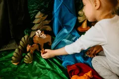 A child kneeling on green silk places a small wooden deer beside a stag under a wooden pine tree, with a blue silk river behind