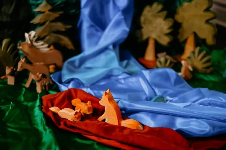 Three wooden fox figures on a red silk beside a blue silk river, with wooden deer and trees on green silk behind them